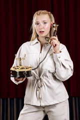 A young, beautiful woman holds an old desk telephone. Close-up against a theater curtain.