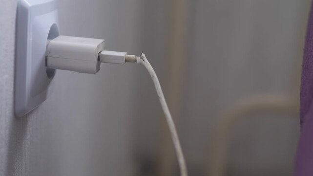  close-up of a man's hand plugging a faulty electrical appliance with a damaged wire into a socket, phone charger with faulty wire connected to power network