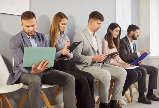 Group of job seekers sitting in row with laptops and papers, waiting for job interview, check their resumes before turn, wanting to get position, dressed formally. Human resources