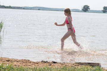 Young girl in pink swimwear joyfully splashing through shallow water at a serene lake, surrounded by lush greenery and a bright sunny sky, capturing the essence of summer fun