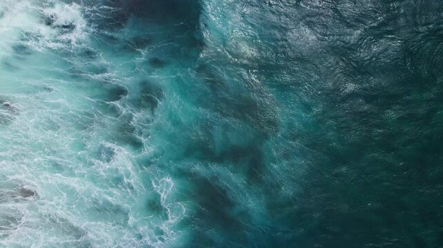 Aerial top-down view of turquoise ocean waves gently rolling across the surface. The foam from the white water contrasts beautifully with the deep blue sea, creating an abstract, soothing pattern. The