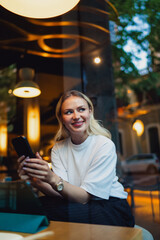 Woman seated by window, smiling with smartphone in both hands