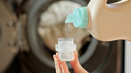 A woman pouring liquid laundry detergent into a cap. Close-up of a woman's hands holding a measuring cap and pouring laundry gel, with a washing machine in the background. Laundry Detergent concept.