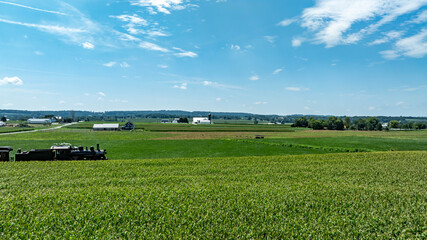 Lush green fields stretch across the landscape, dotted with farm buildings and equipment. The scene captures the tranquility of rural life on a sunny day in the countryside.