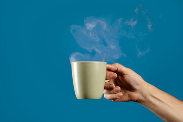 Hand holding steaming coffee mug on blue background. Man enjoying hot drink like tea or coffee,...