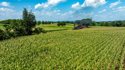 A vintage steam train moves slowly through green cornfields on a sunny day. The landscape features blue skies dotted with clouds, creating a peaceful rural scene.