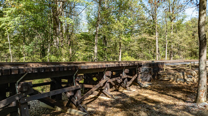 A weathered wooden railway bridge stands elevated over a quiet landscape. Tall trees surround the structure, showcasing vibrant greenery, while sunlight filters through the leaves.