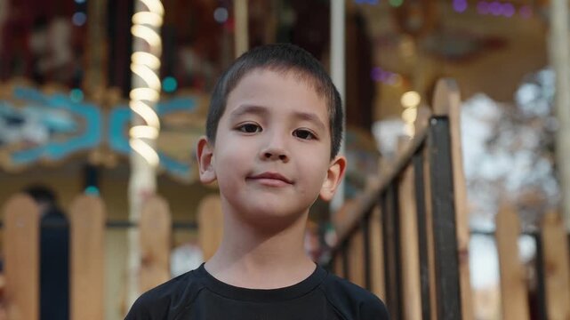 Close-up portrait of a young boy with a confident smile standing outdoors near a carousel. Warm daylight, soft bokeh, real lifestyle moment with natural emotion.