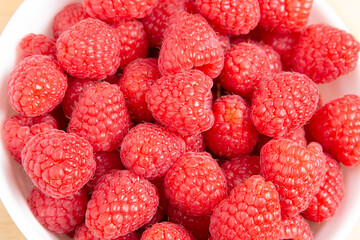 Extreme close-up of a white bowl overflowing with ripe red raspberries on a wood surface

