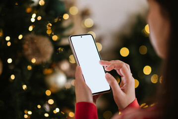 Woman using a smartphone with a blank screen near a Christmas tree decorated with warm festive lights.
