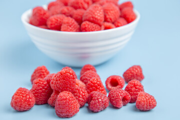Ripe red raspberries in the foreground, with a blurry white bowl of fruit behind on blue