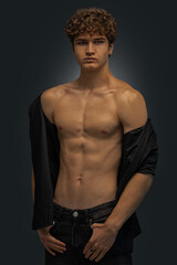 Fototapeta premium young man stands in a studio showcasing his curly hair and toned physique. He wears a black shirt casually draped over one shoulder while looking directly at the camera.