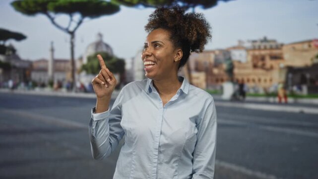 Woman with curly hair points finger upward and smiles broadly while standing on a city street lined with historic buildings and pedestrians; joy curiosity discovery.