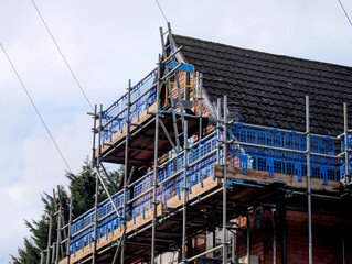 Residential brick house roof under renovation with blue safety scaffolding against a bright sky in England
