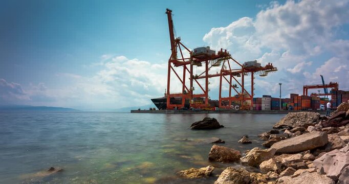 Rijeka, Croatia - August 21, 2025: Timelapse. Container port with large orange cranes rising above the calm blue sea, viewed from a rocky shoreline under a summer sky.