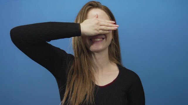 Woman covers eyes with hand against bright blue studio wall hiding expression; privacy mystery introspection shyness.