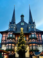 Half-timbered Wernigerode Town Hall, Wernigerode, Germany, features two pointed towers against a blue sky. A tall, illuminated Christmas tree 