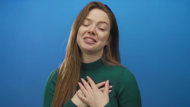 Young caucasian woman softly places hand on chest in studio setting against blue background; gratitude.