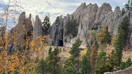 Needles Highway shows road, tunnel & Black Hills in South Dakota. Pointed mountains look like needles. Road curves as auto leaves one of many tunnels. Road is for auto tours. Trees show fall yellow.