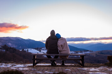 Couple in love enjoying landscape of winter Carpathian mountains sitting on bench at sunset. Back view. Space