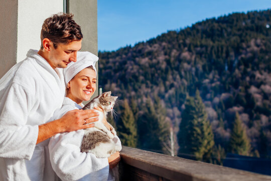 Young couple holding cat on hotel balcony enjoying winter mountain landscape wearing bathrobes. Travel with pet