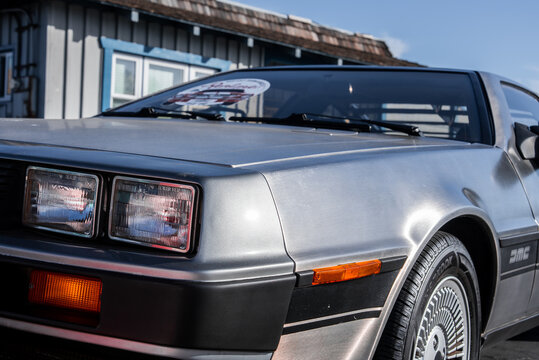 Stainless steel DeLorean DMC 12 parked by a coastal style building in Monterey, California. Square headlights, amber marker, turbine wheel, and DMC badge show in crisp daylight.