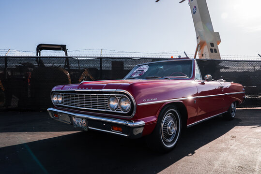 A red 1964 Chevrolet Chevelle convertible sits by a barbed fence in Monterey, with a crane and equipment behind. Afternoon light adds strong contrast and long shadows.