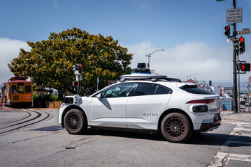 Obraz premium A white Waymo SUV with lidar and sensors waits at a light by cable car tracks in San Francisco, near Hyde Street and Beach Street, in cool midday foggy light.