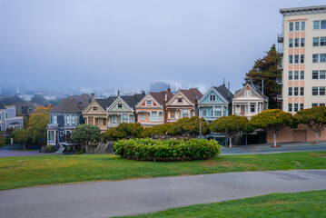Colorful Victorian houses at Alamo Square in San Francisco stand by a park lawn and hedge as fog softens the distant skyline, suggesting a cool overcast day.