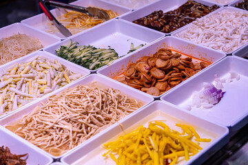 A close-up of a vibrant Chinese cold dish buffet with sliced meats, noodles, shredded vegetables (cucumber, tofu), and bamboo shoots. The display highlights Asian cuisine variety.
