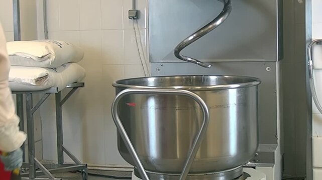 A worker in a red apron and gloves prepares a large industrial mixer in a food production facility. The mixer is stainless steel. Bags of ingredients wait to be added to the mix. It's daytime.