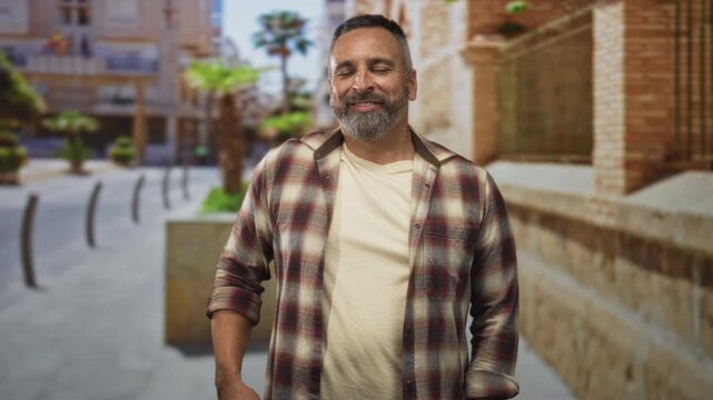 Middle age man with grey beard smiling with eyes half closed, standing on a sunny street plaza with hands relaxed at sides; contentment confidence.