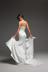 Naklejka premium Elegant bride poses in minimalist white satin wedding gown against soft natural light backdrop