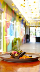 Platillo gourmet mexicano con tortilla oscura, decorado con jalapeño, cebolla encurtida y crema amarilla sobre pétalos de flor.