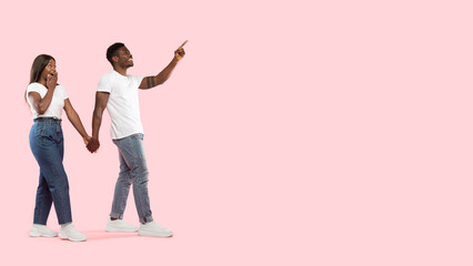 Fototapeta premium A joyful black couple walks hand in hand, with the man pointing excitedly to the side. The backdrop is a bright pink studio, capturing their happy expression and energy.