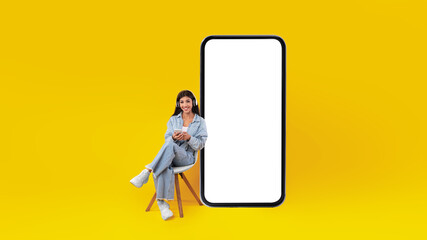 A young woman is sitting comfortably on a chair, holding a smartphone. Beside her is a large blank...
