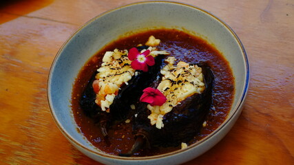 Platillo gourmet con chiles rellenos bañados en salsa roja, con camarón y decorados con pétalos comestibles.