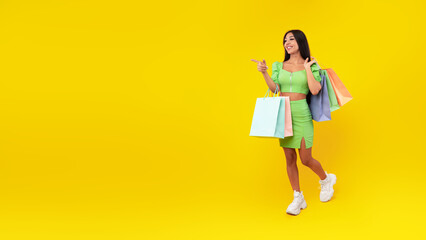 A young woman in a stylish green outfit smiles while walking and pointing. She holds multiple shopping bags in soft pastel colors. The bright yellow background creates a cheerful mood.