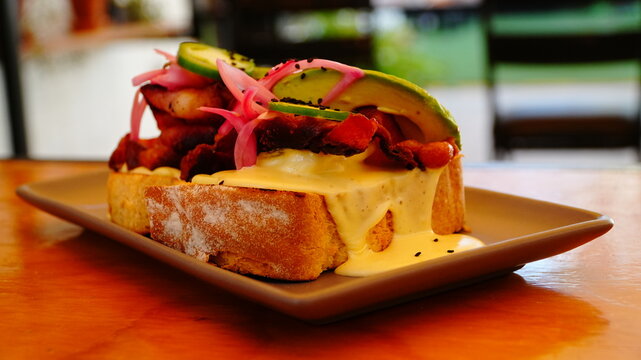 Tostada gourmet con pan artesanal, ba&ntilde;ada en cremosa salsa de queso y coronada con tiras de tocino crujiente, aguacate fresco y cebolla morada encurtida.