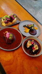 cuatro platillos gourmet mexicanos sobre mesa de madera: tostadas con aguacate y tocino, tortilla negra decorada, chiles rellenos en salsa y costilla en adobo.