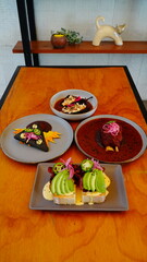 cuatro platillos gourmet mexicanos sobre mesa de madera: tostadas con aguacate y tocino, tortilla negra decorada, chiles rellenos en salsa y costilla en adobo.