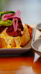Tostada gourmet con pan artesanal, bañada en cremosa salsa con huevo y coronada con tiras de tocino crujiente, aguacate fresco y cebolla morada encurtida.