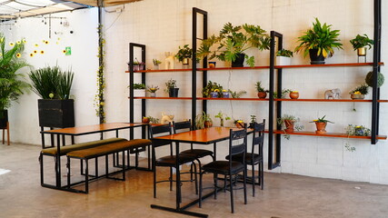 Terraza luminosa y acogedora con mesas de madera y sillas negras, decorada con plantas naturales y estanterías verdes. El espacio cuenta con toldos que brindan sombra y un ambiente relajado