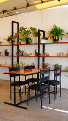 Terraza luminosa y acogedora con mesas de madera y sillas negras, decorada con plantas naturales y estanterías verdes. El espacio cuenta con toldos que brindan sombra y un ambiente relajado