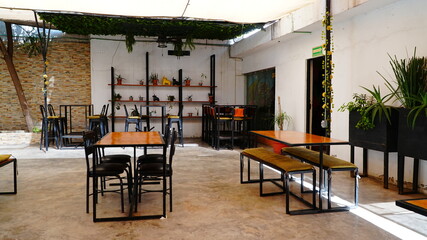 Terraza luminosa y acogedora con mesas de madera y sillas negras, decorada con plantas naturales y estanterías verdes. El espacio cuenta con toldos que brindan sombra y un ambiente relajado