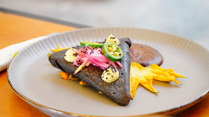Platillo gourmet mexicano con tortilla oscura, decorado con jalapeño, cebolla encurtida y crema amarilla sobre pétalos de flor.