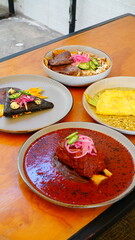 Cuatro platillos mexicanos servidos sobre una mesa de madera, cada uno con presentaciones coloridas y cuidadas: carne en salsa roja, tortilla negra, omelette con flor de calabaza y chiles rellenos.