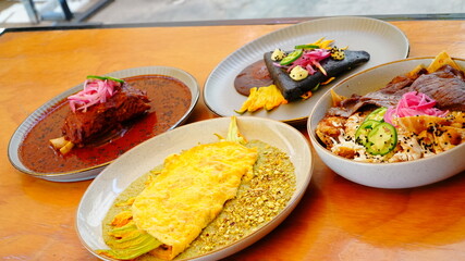Cuatro platillos mexicanos servidos sobre una mesa de madera, cada uno con presentaciones coloridas y cuidadas: carne en salsa roja, tortilla negra, omelette con flor de calabaza y chiles rellenos.