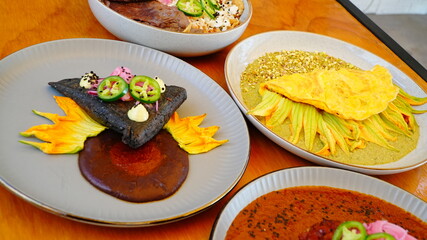 Cuatro platillos mexicanos servidos sobre una mesa de madera, cada uno con presentaciones coloridas y cuidadas: carne en salsa roja, tortilla negra, omelette con flor de calabaza y chiles rellenos.