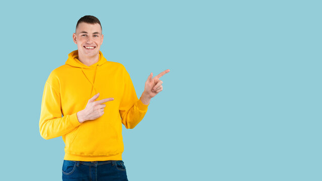A young man with short hair smiles while pointing to the right. He is wearing a bright yellow hoodie and blue jeans against a light blue background, creating a cheerful atmosphere.